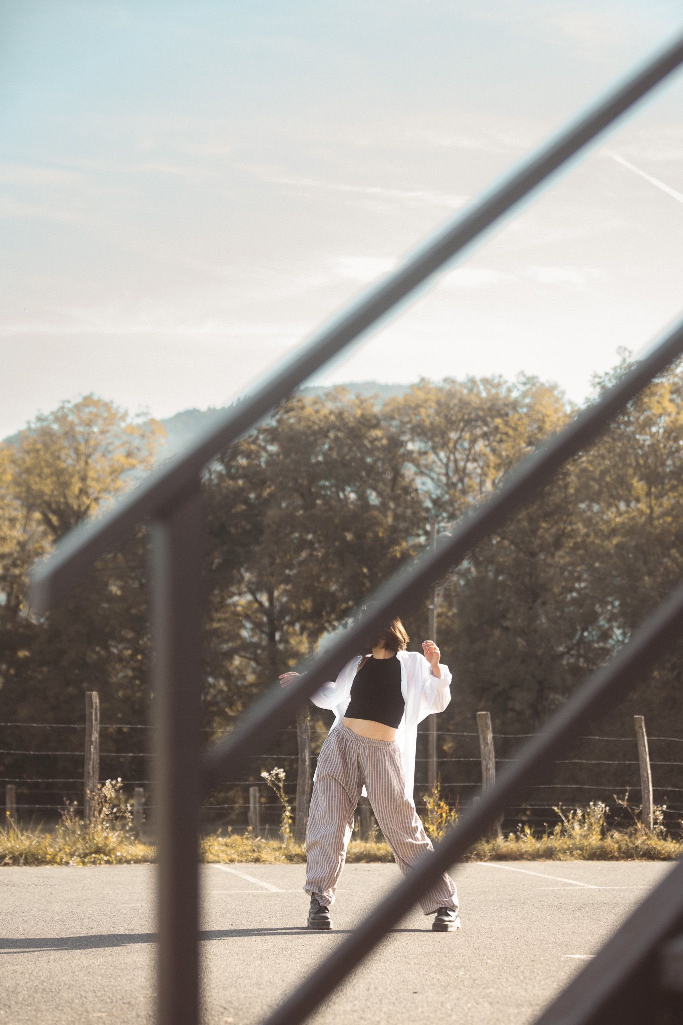 Female dancer in white outfit performing outdoors on a court viewed through a metal fence.