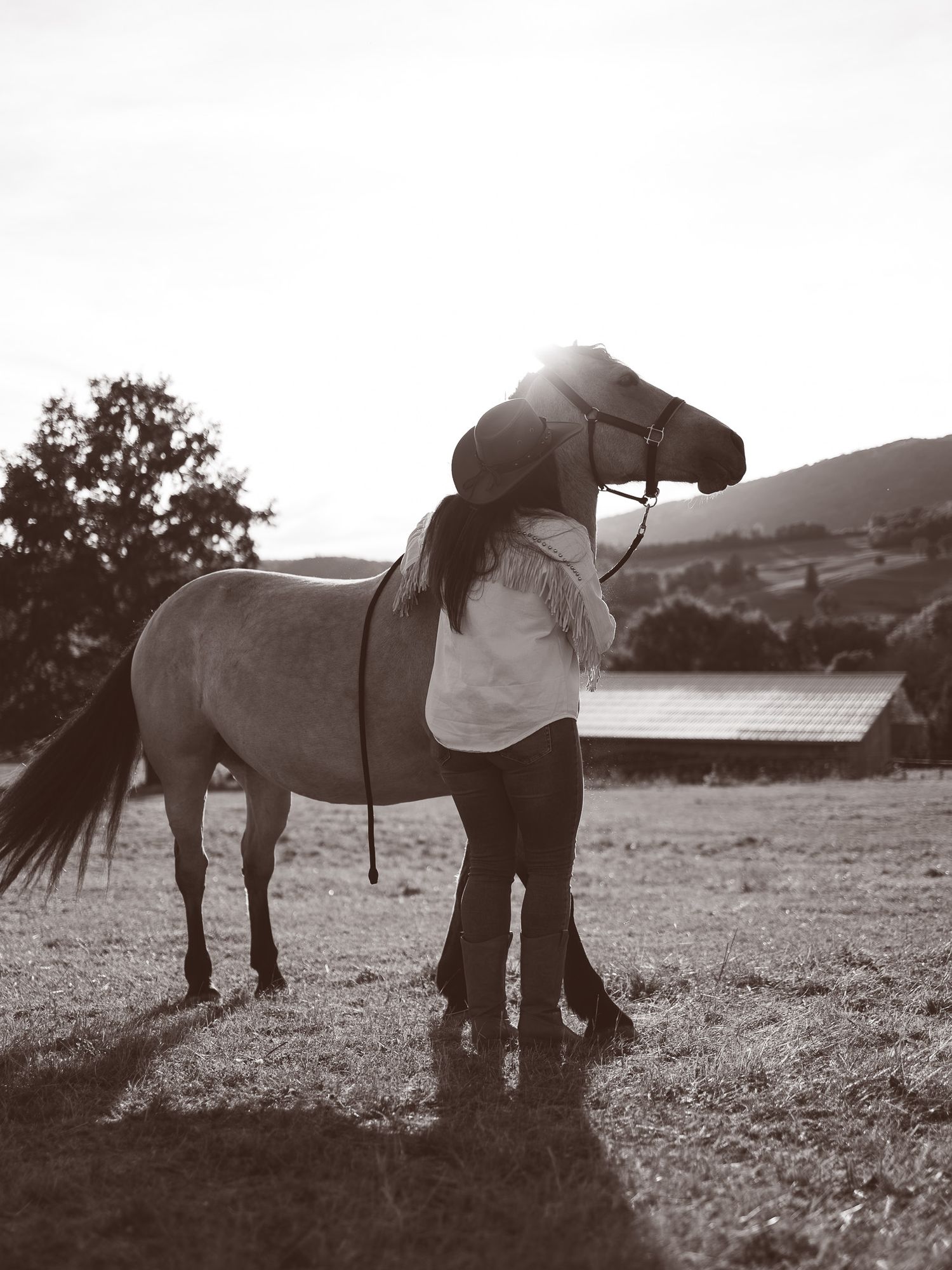 Black and white photo of woman in cowboy hat embracing white horse in rural field at sunset.