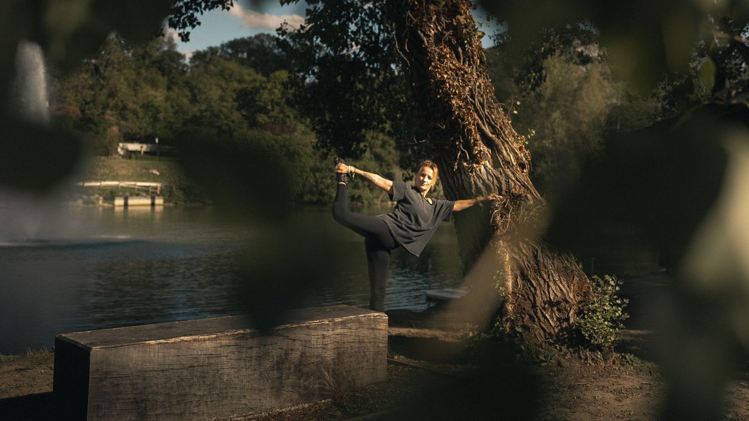 A dancer performs a high kick near a large tree beside a calm river at dusk.