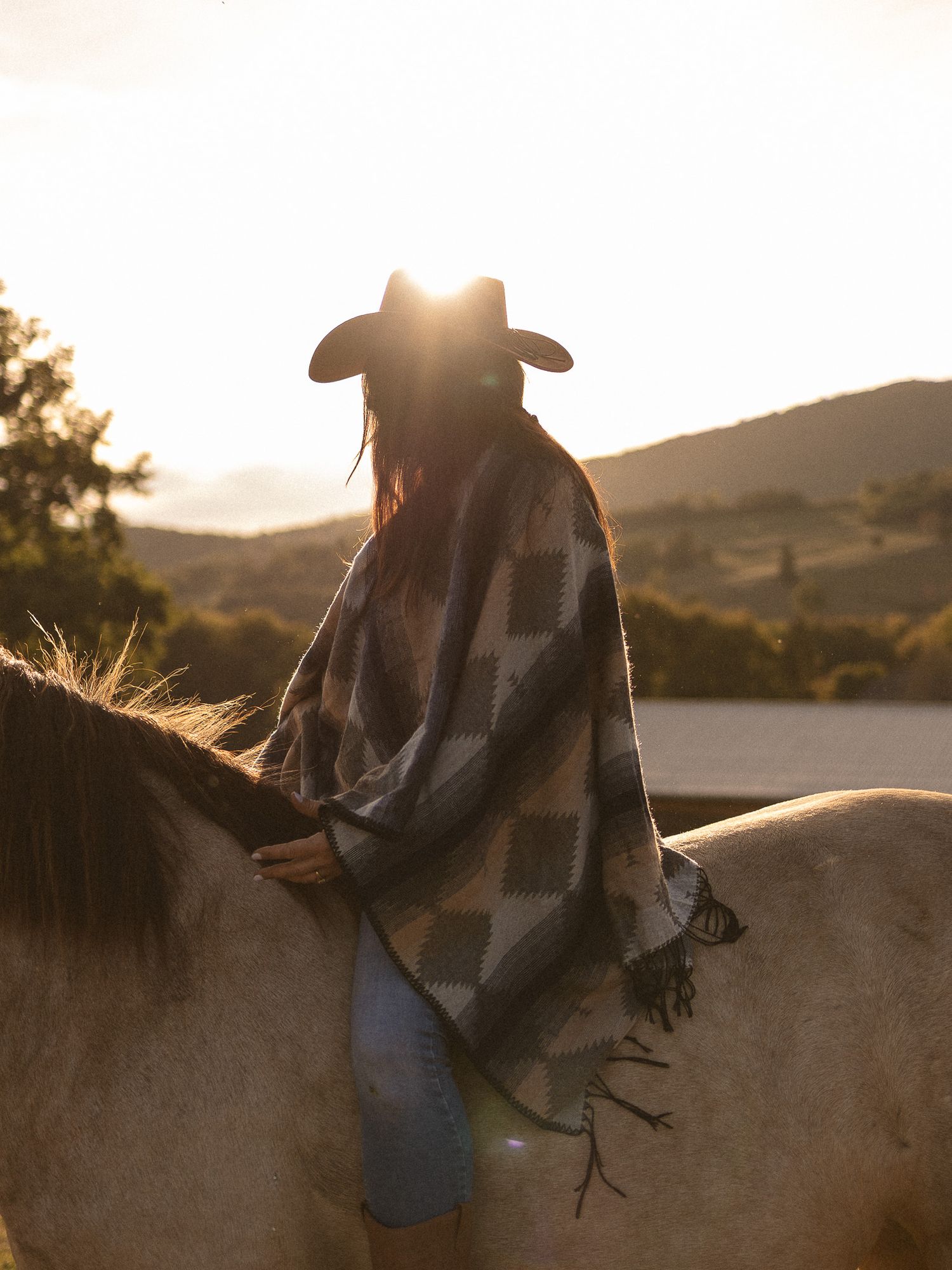 Woman in cowboy hat and patterned poncho riding a white horse at sunset with scenic hills.