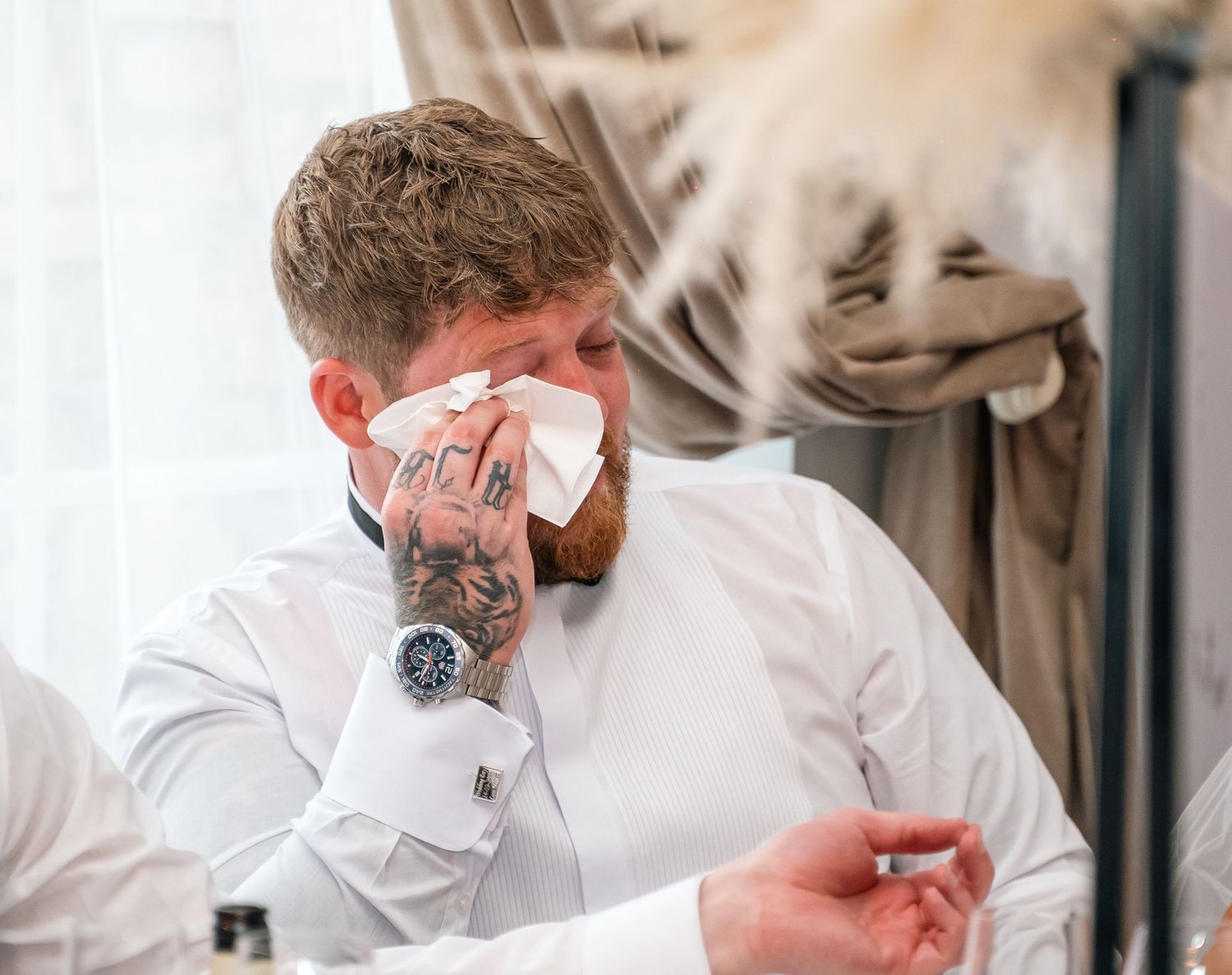 Emotional tattooed groom wiping tears with a tissue during wedding ceremony.