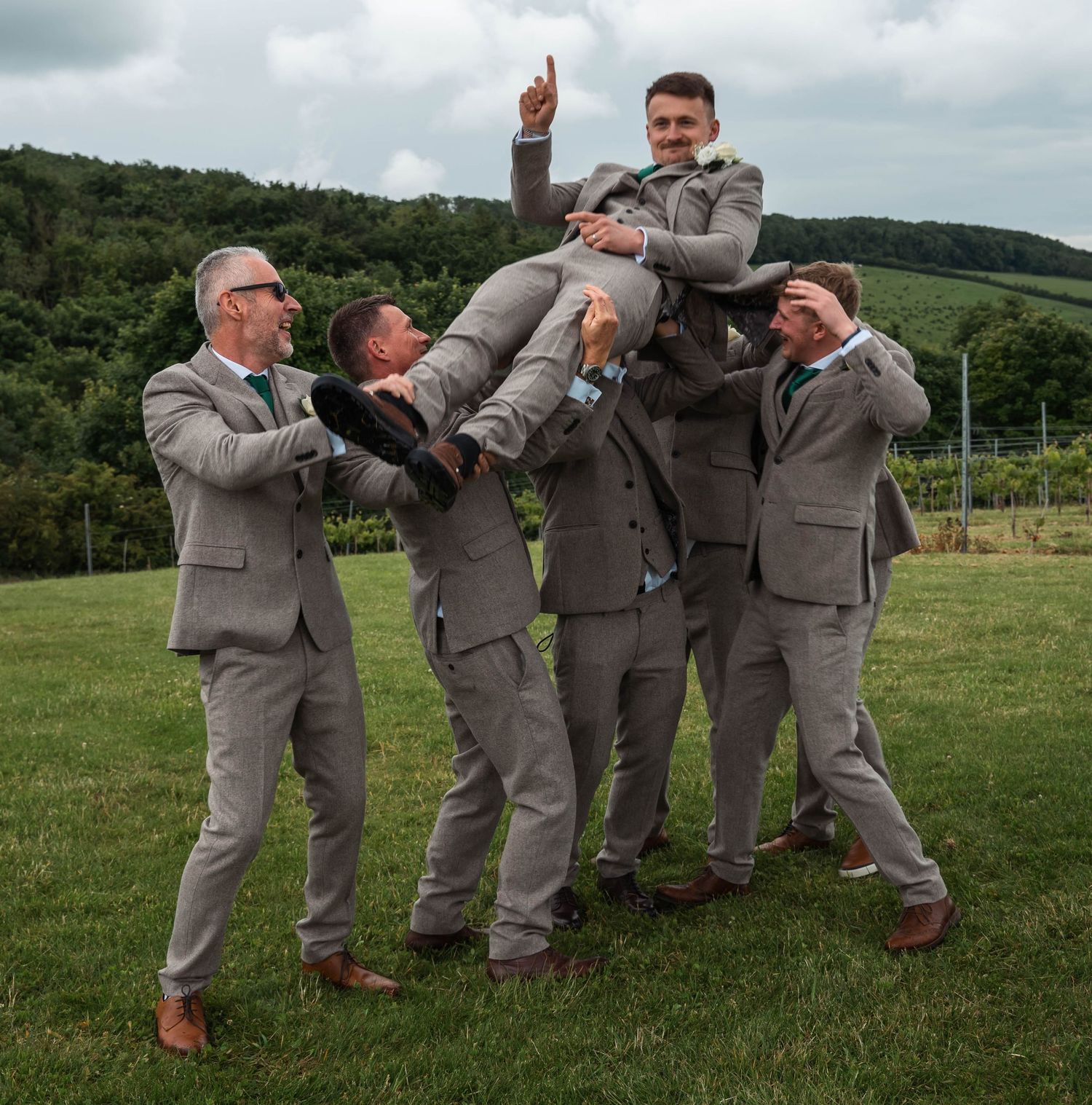 Groomsmen lift celebrating groom into the air outdoors at vineyard wedding venue.