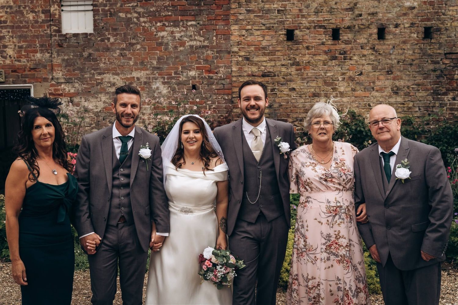 Wedding party group photo with bride, groom, and family members outside a brick venue.