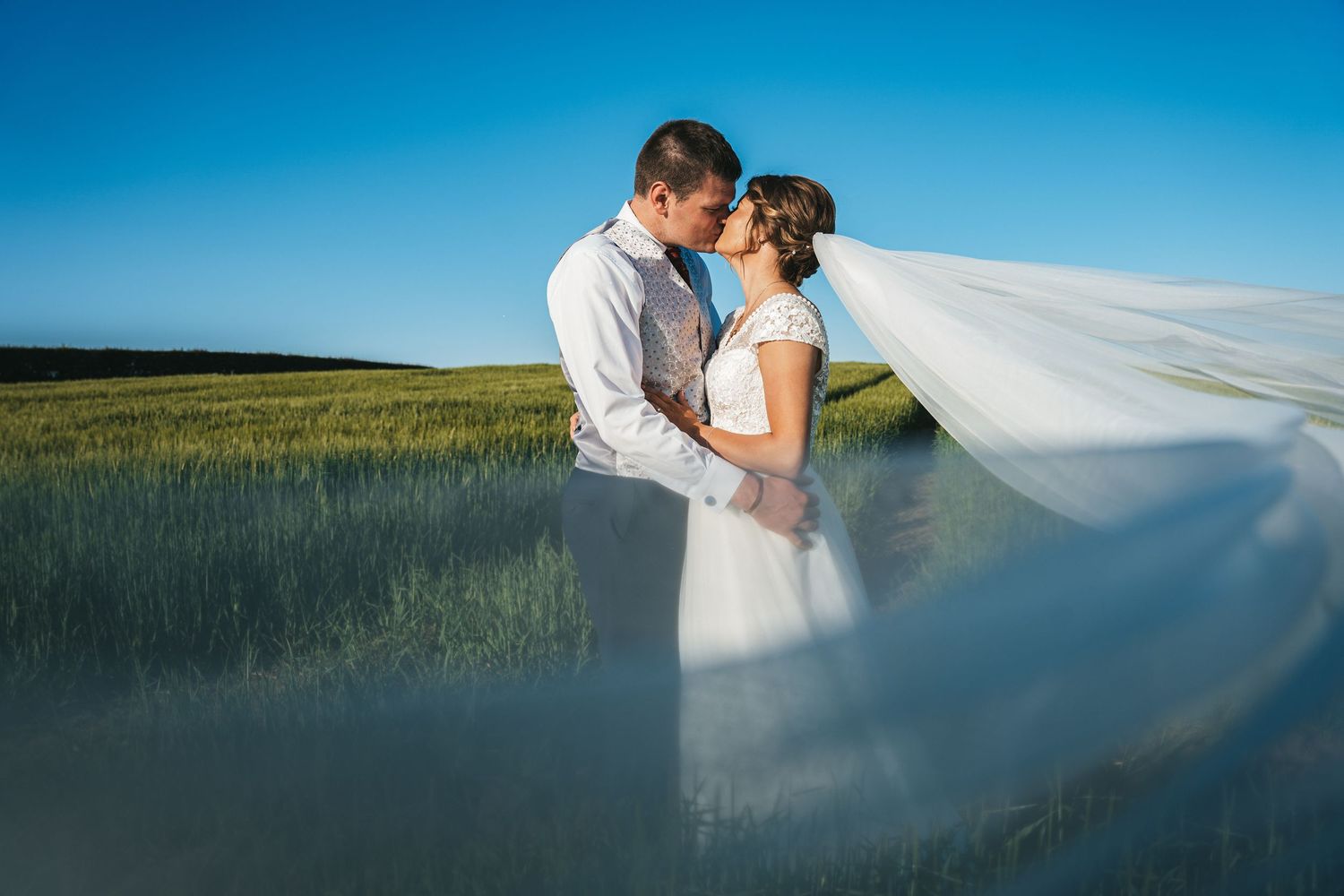 Bride and groom kissing in a green field with wedding veil flowing in the wind.