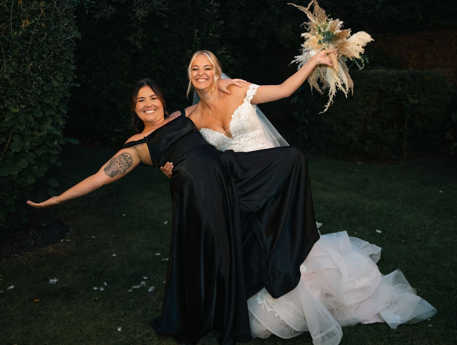 Bride in white ruffled gown and bridesmaid in black dress joyfully pose in garden.