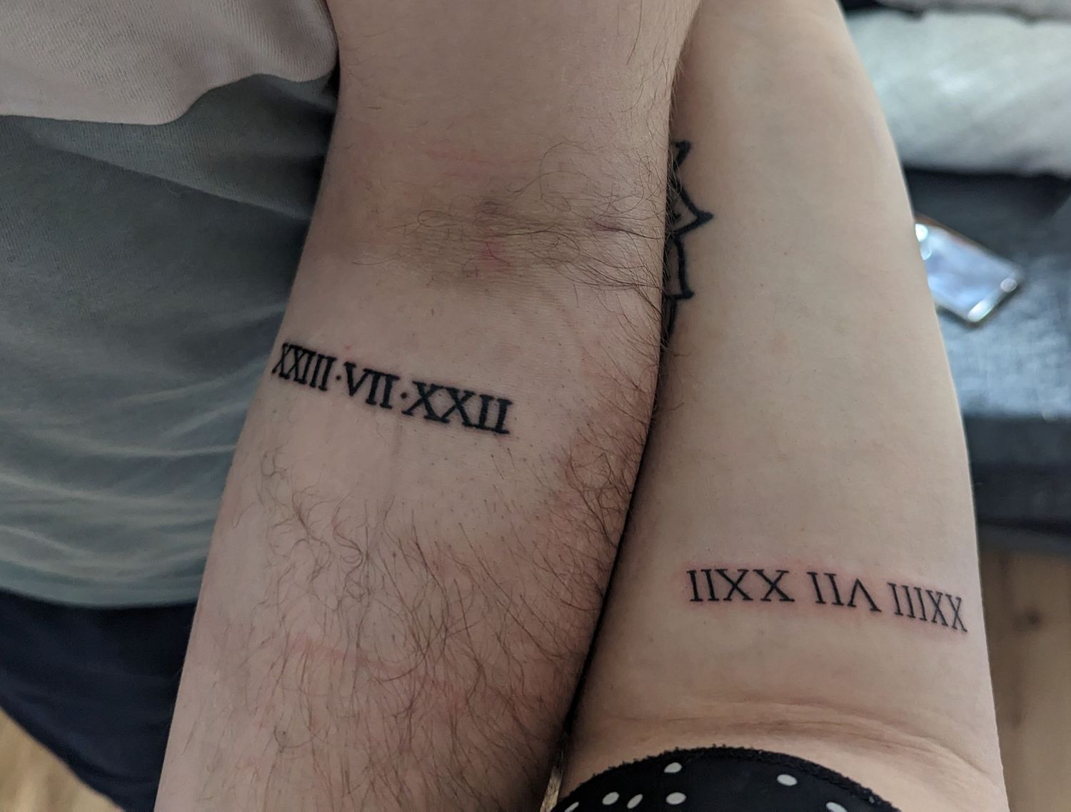 Two arms side by side displaying matching Roman numeral date tattoos on inner forearms.