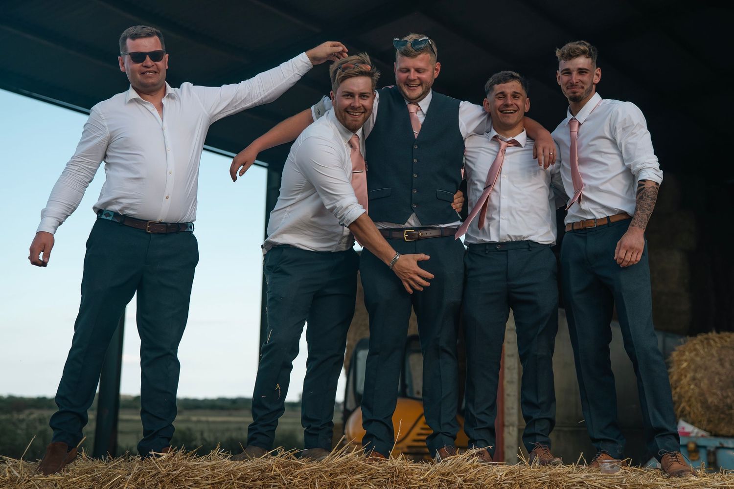 Five groomsmen laughing and posing together in front of hay bales at a rustic outdoor wedding reception.