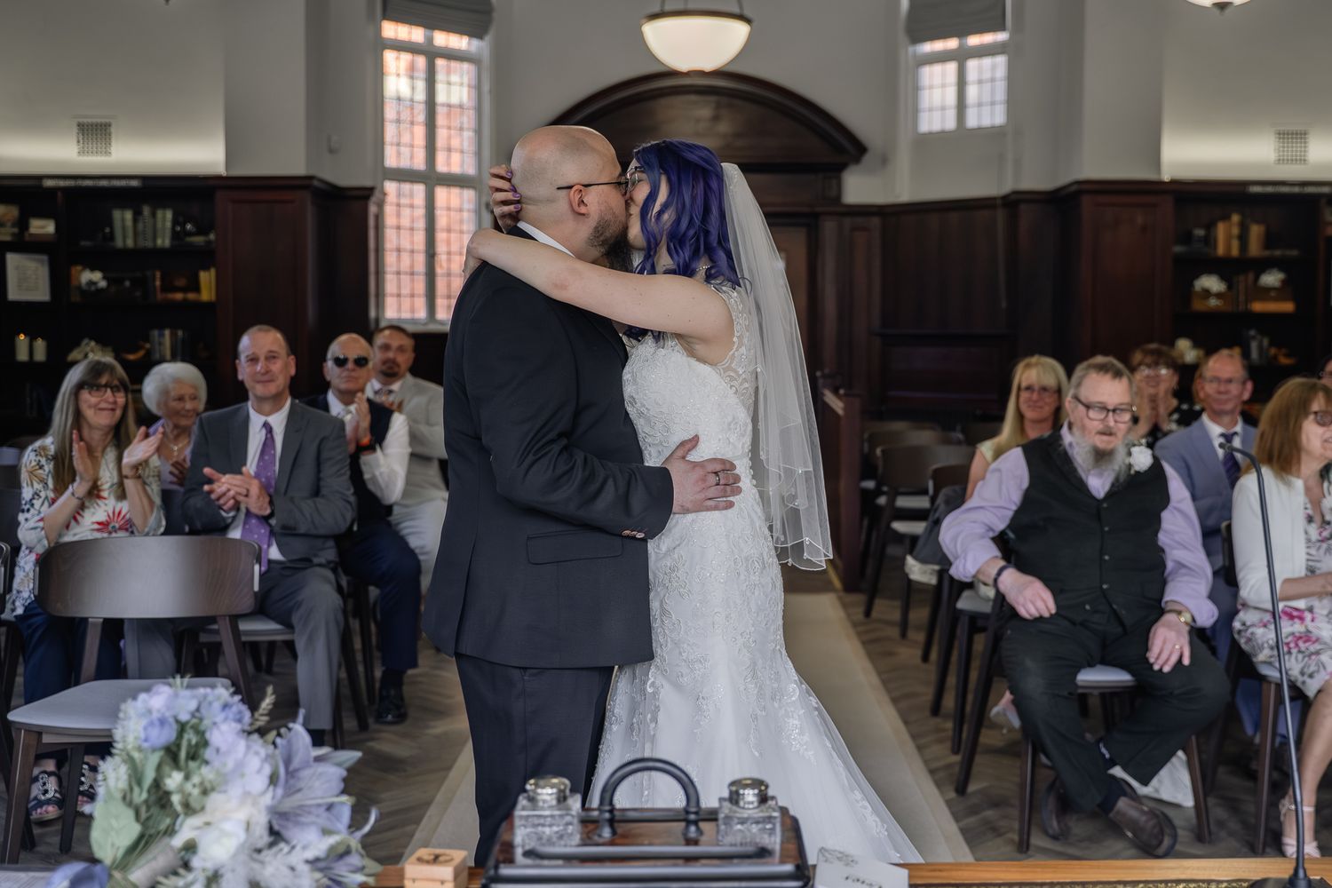 Newlyweds sharing their first kiss at a wedding ceremony while guests applaud in background.