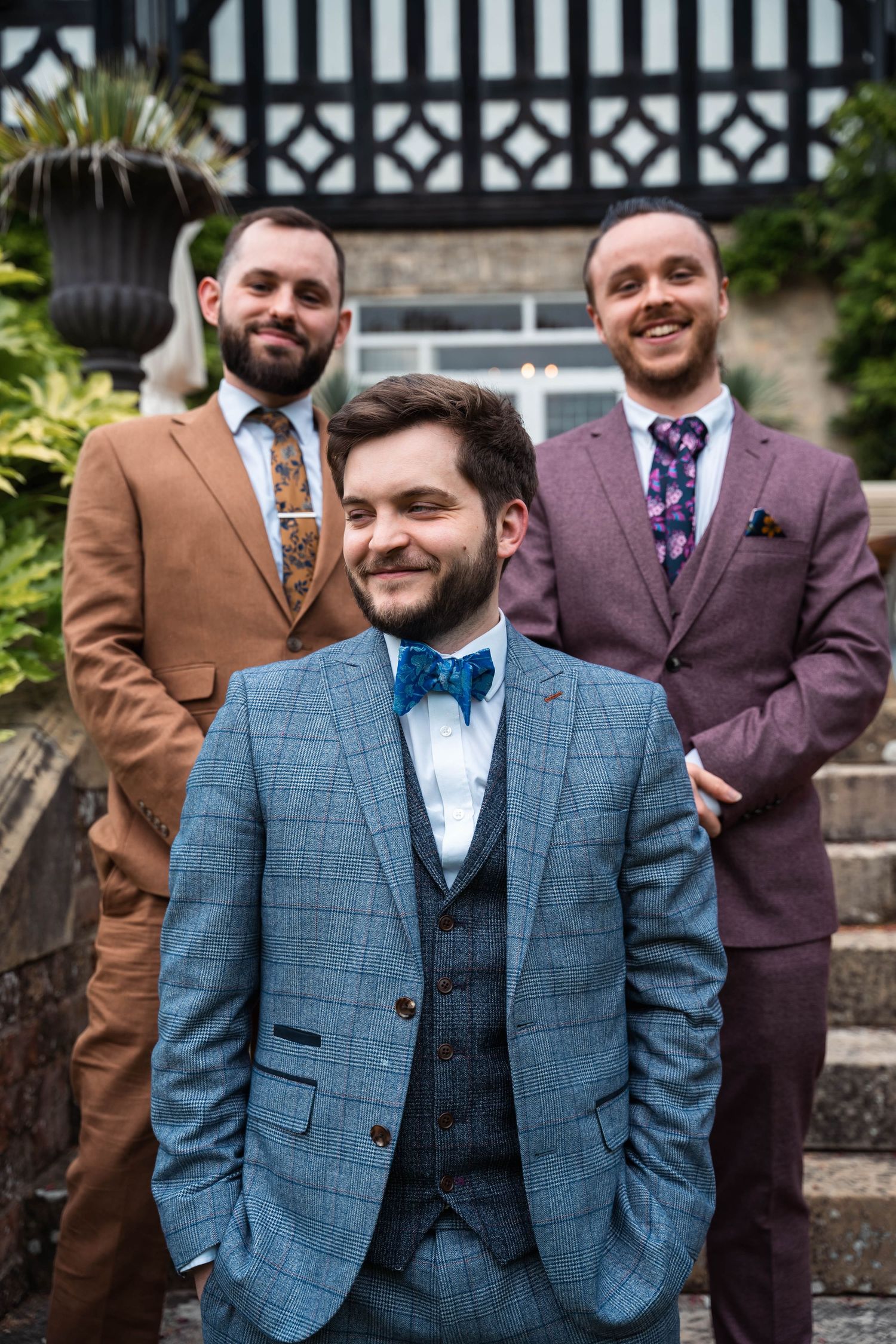 Groom in blue suit with two groomsmen in colorful suits smiling on outdoor steps.