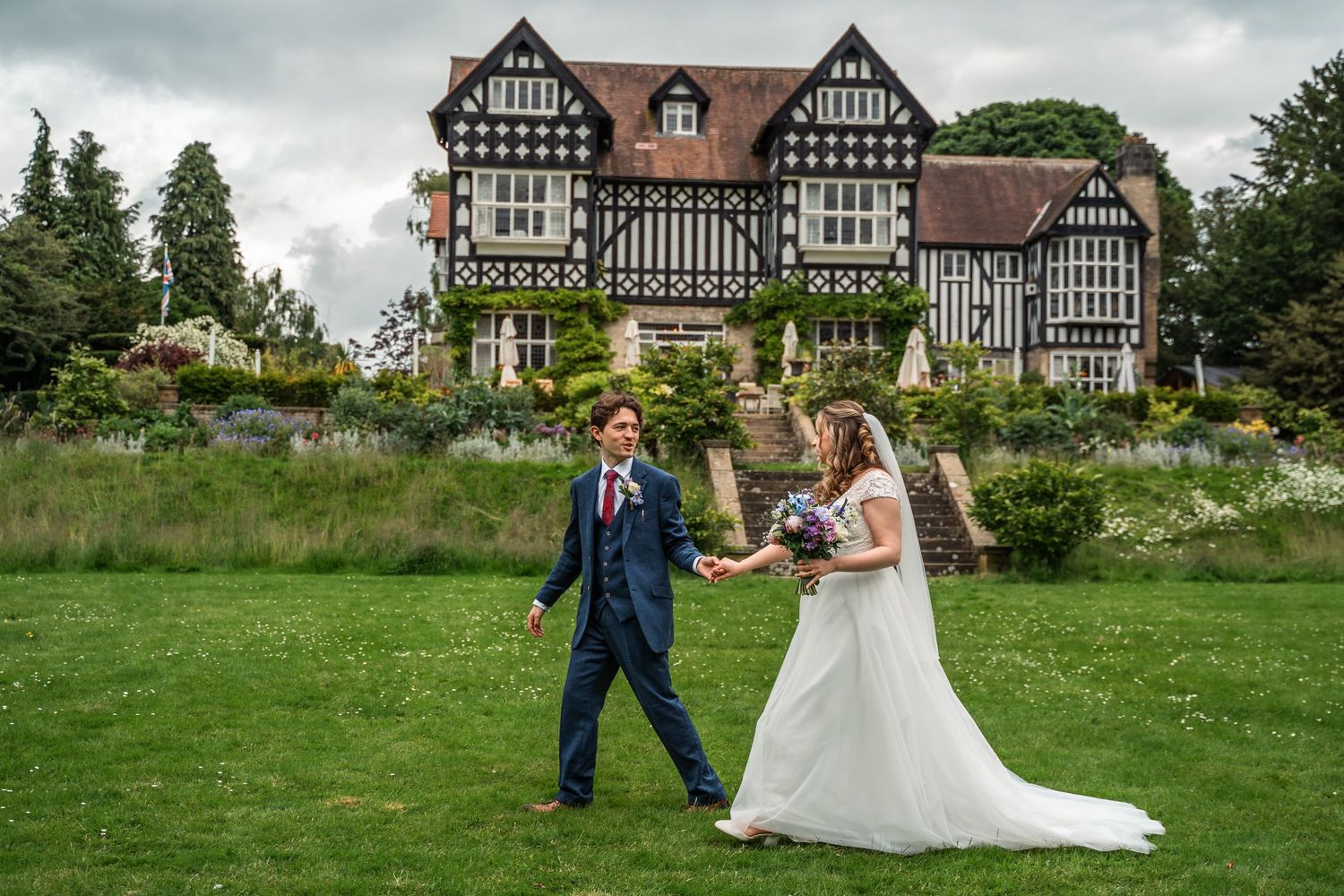 Bride and groom walk hand in hand on lawn before stunning Tudor manor house.