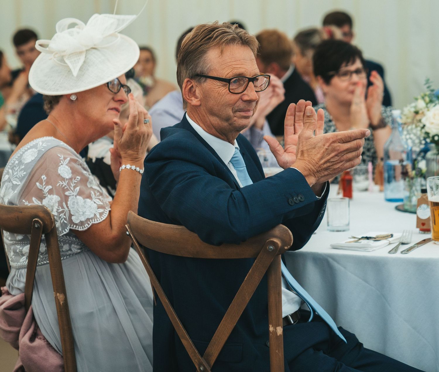 Wedding guests clapping and smiling at a reception dinner under a marquee tent.