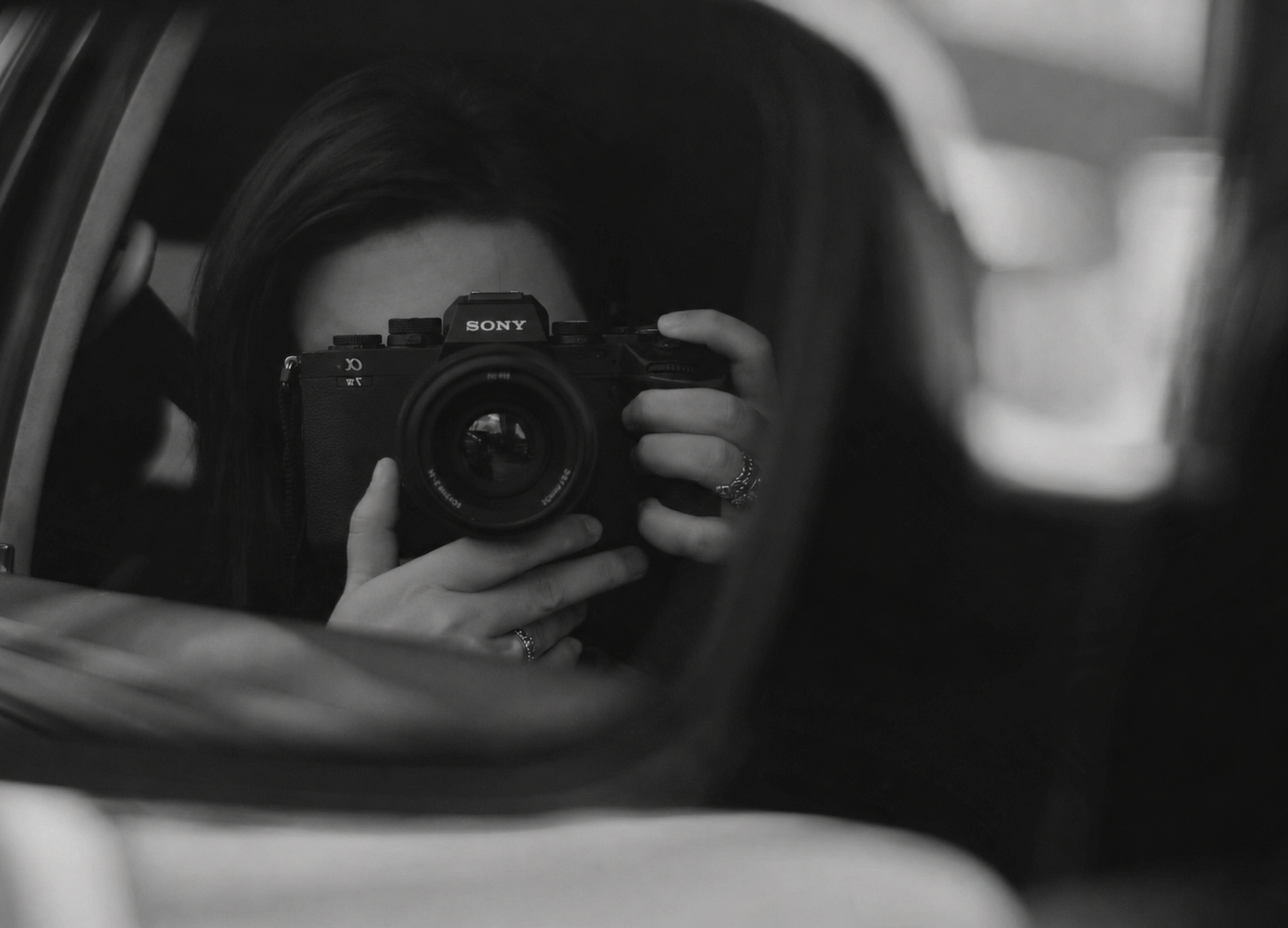 Black and white car mirror selfie of a photographer holding a Sony camera.