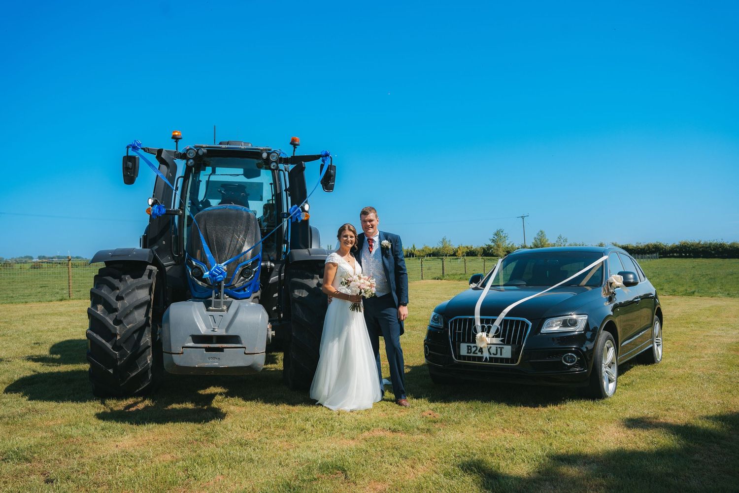 Bride and groom posing between a blue tractor and black Audi on a sunny farm wedding day.