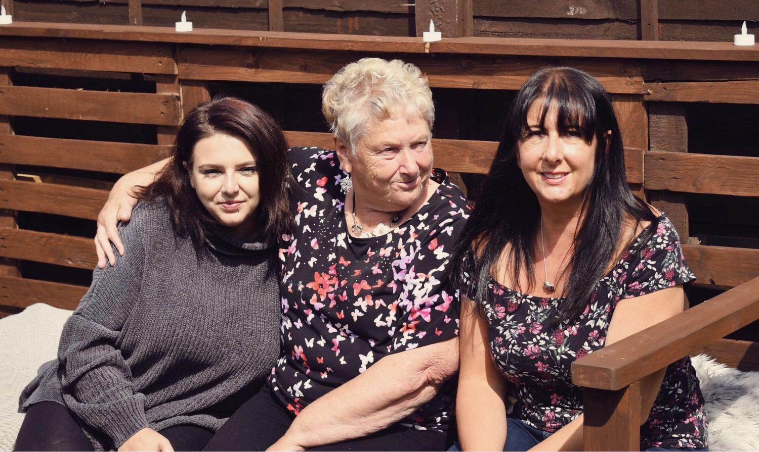 Three women sitting together on a bench outdoors, smiling warmly in casual attire on a sunny day.