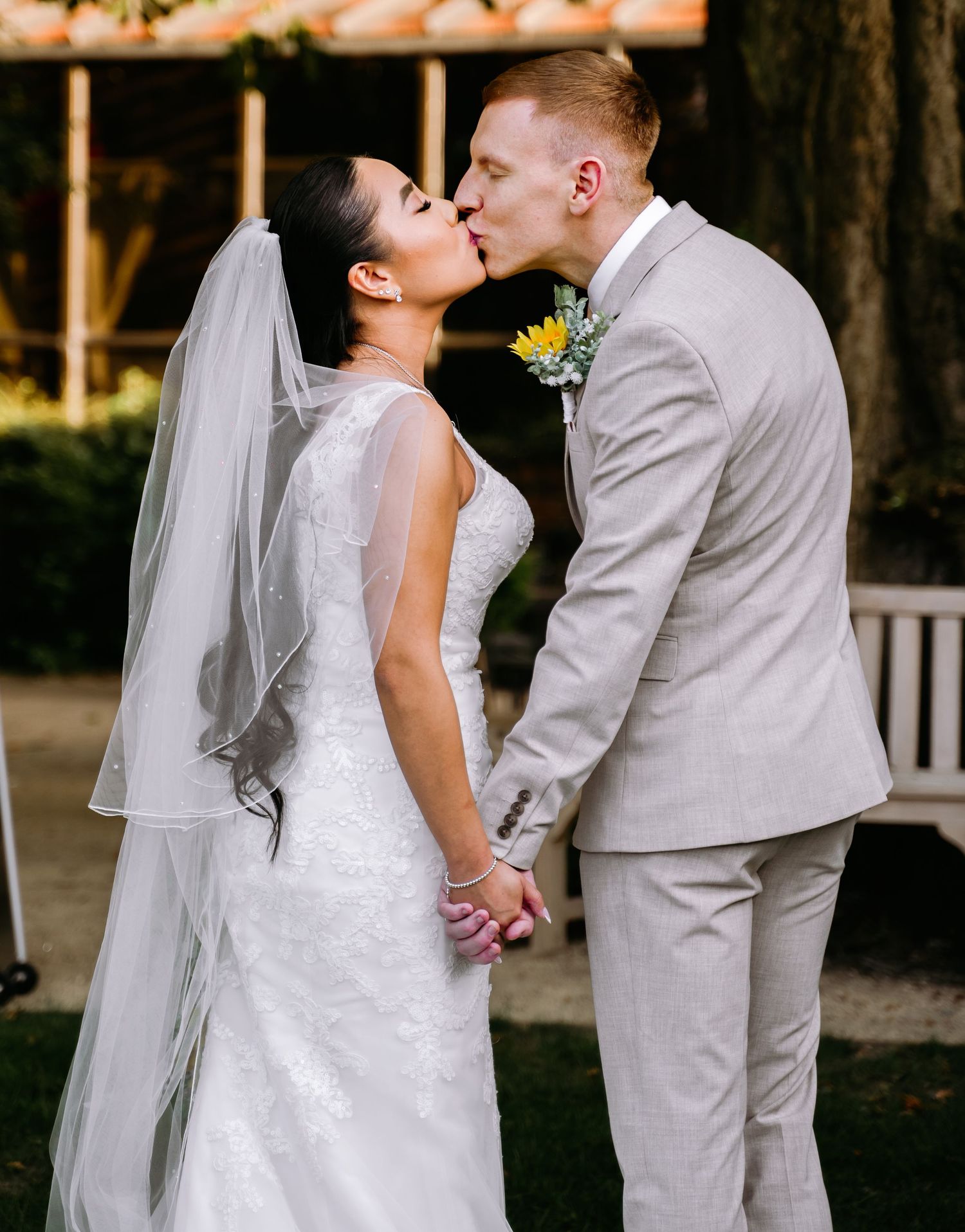 Newlywed couple shares romantic kiss while holding hands outdoors after wedding ceremony.