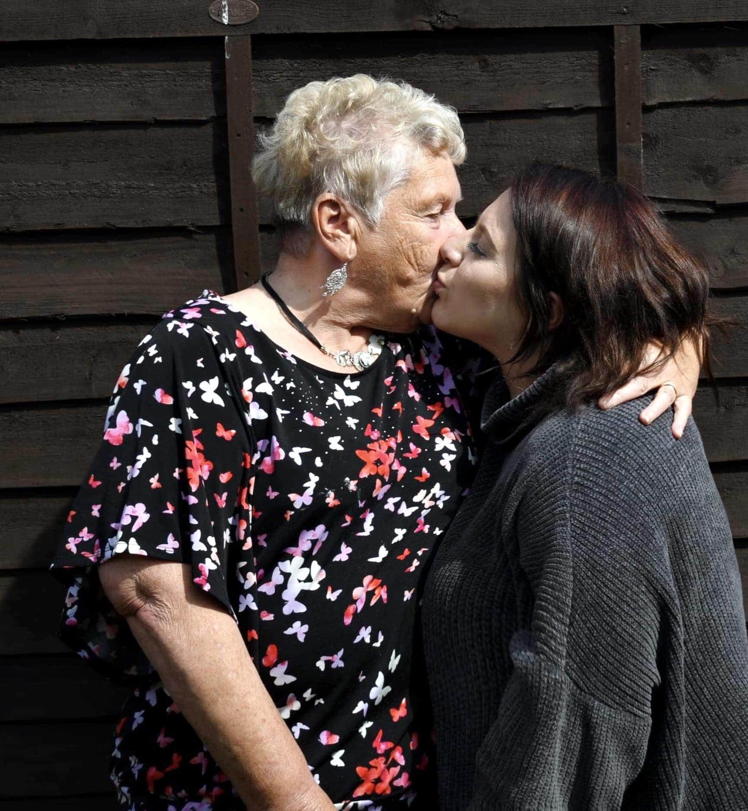 Younger woman with dark hair kissing an older white-haired woman on the cheek outdoors.