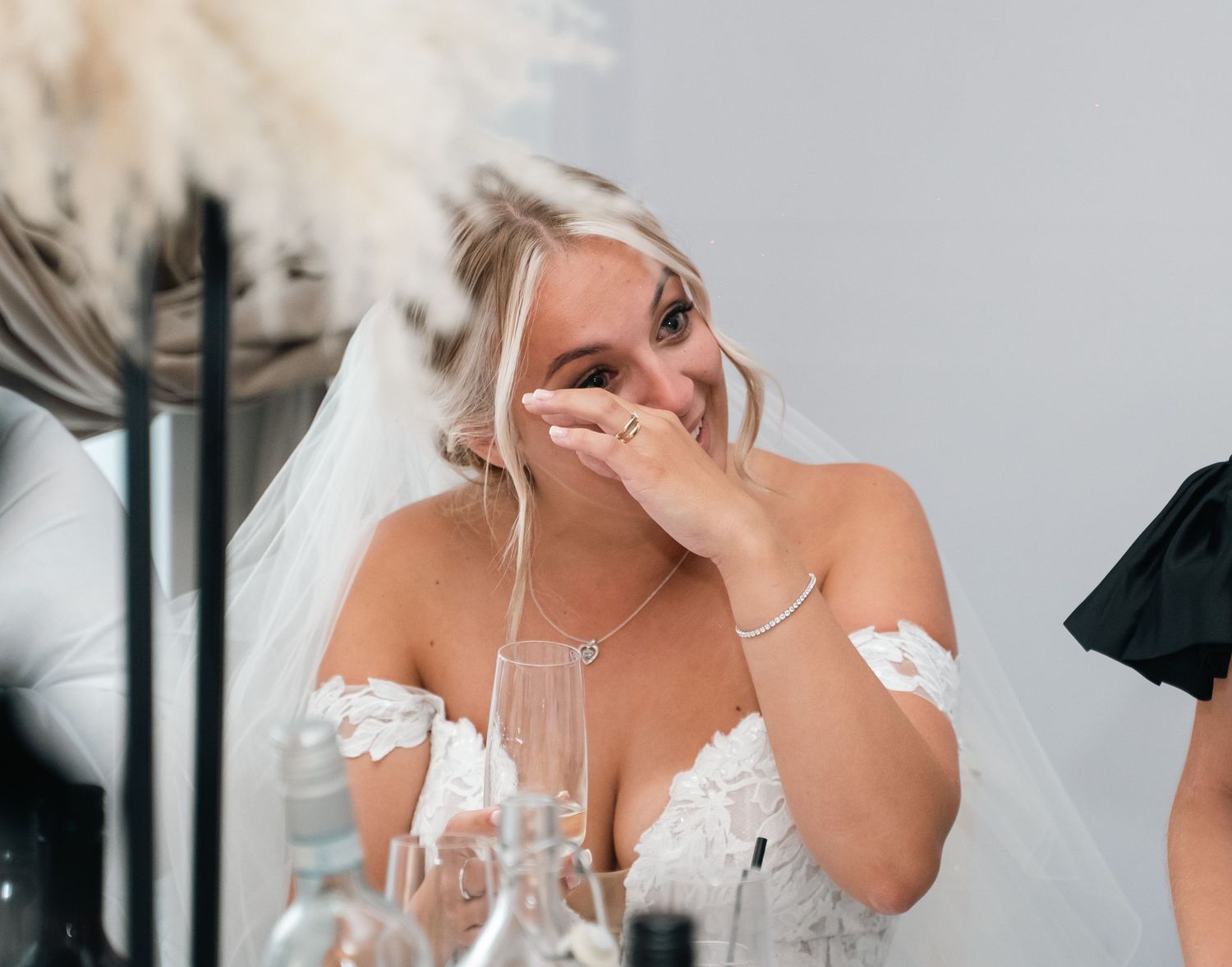 Emotional bride in white veil laughs and covers mouth during wedding reception speeches.