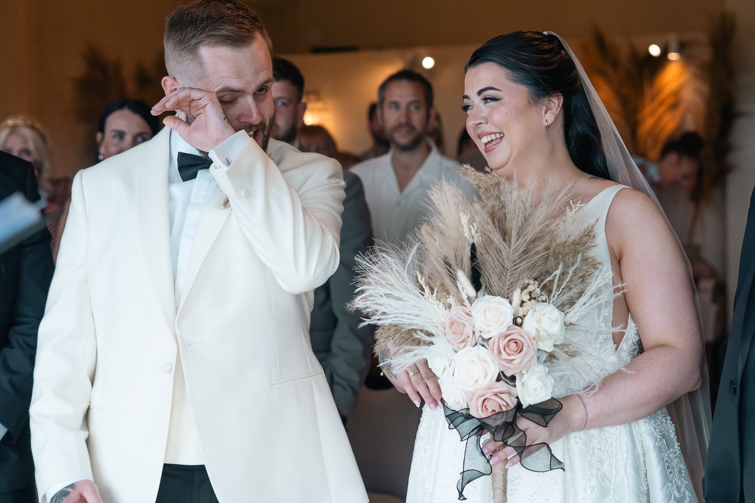 Emotional groom wipes tears while laughing bride holds bouquet during joyful wedding ceremony.