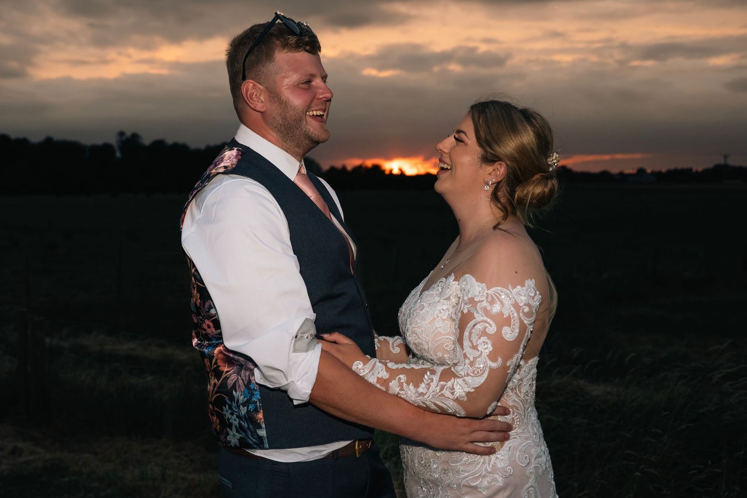 Bride and groom share a joyful moment together in a field during a stunning golden sunset.