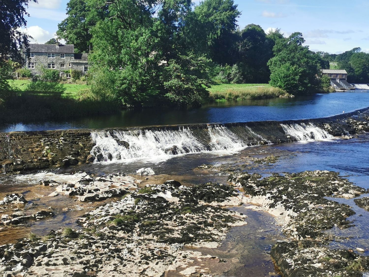 Scenic waterfall cascading over rocks on a river surrounded by lush green trees and buildings.