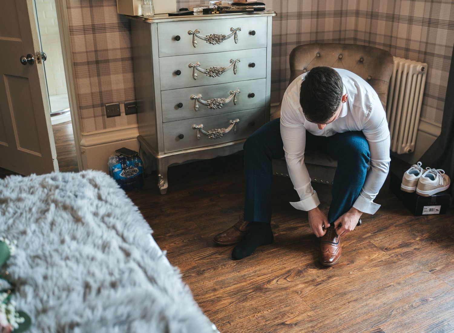 Groom ties his dress shoes while seated in bedroom preparing for wedding day.