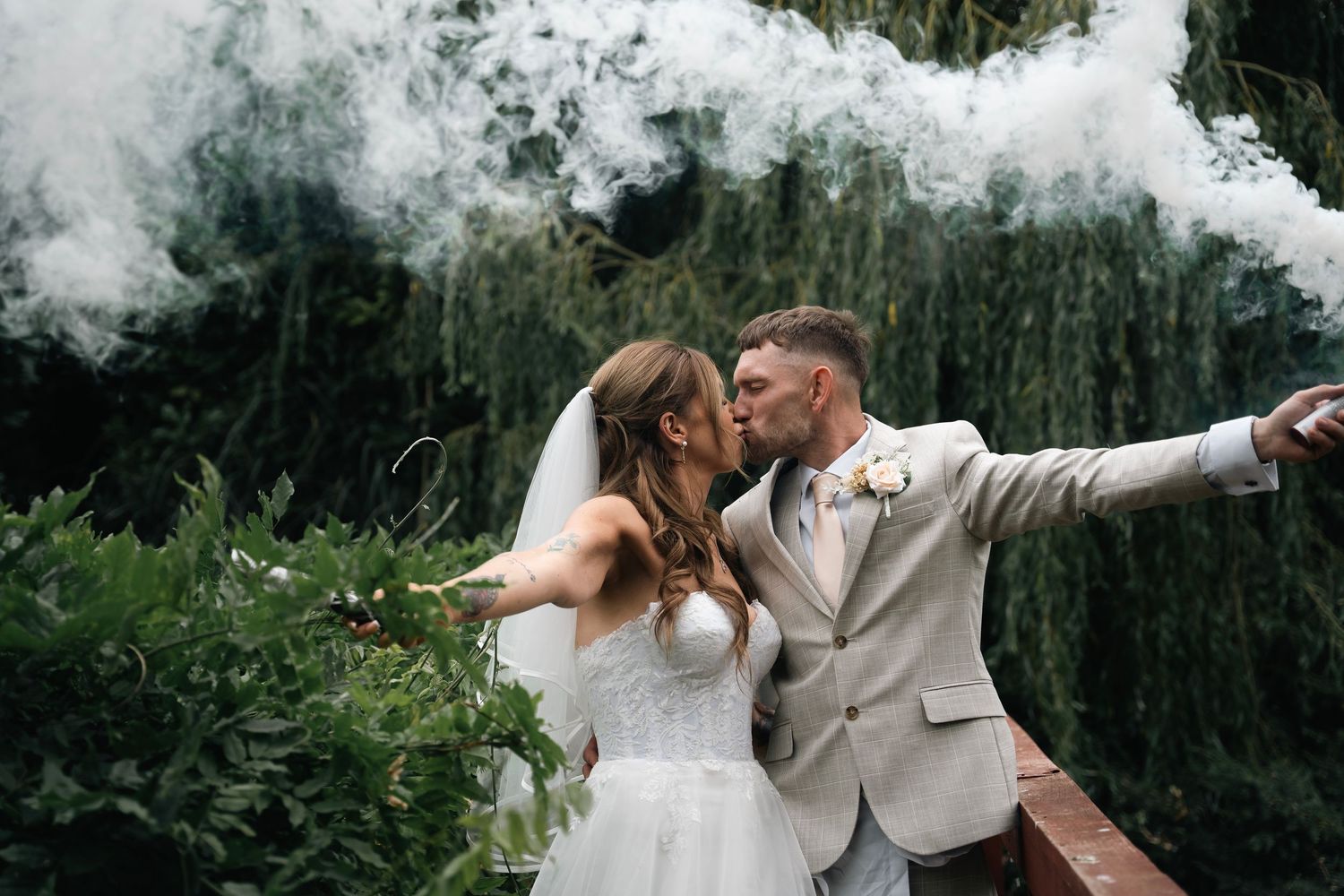 Bride and groom kissing with dramatic smoke bomb effect in garden setting.