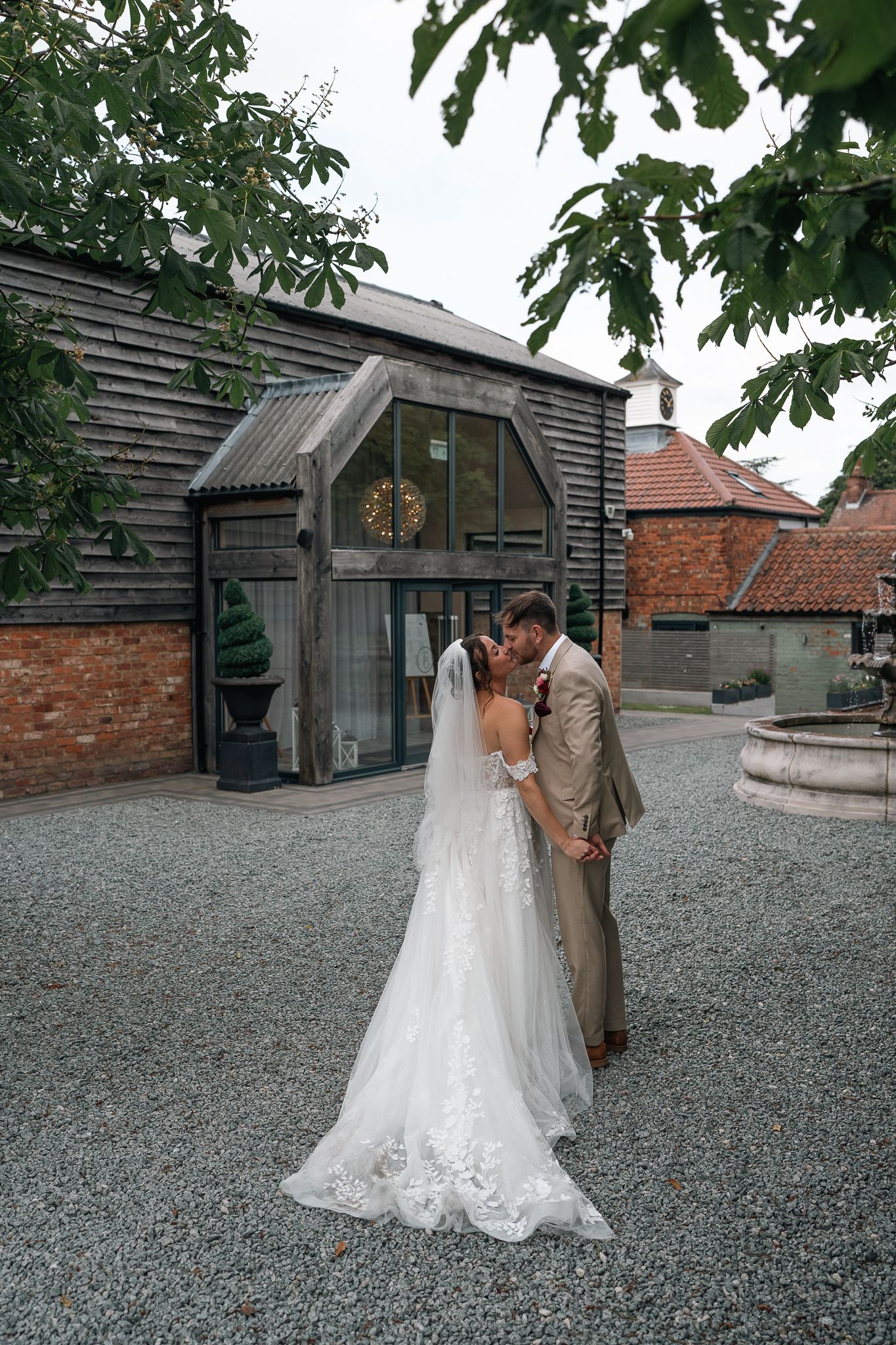 Bride and groom kiss outside rustic barn wedding venue surrounded by lush green trees.