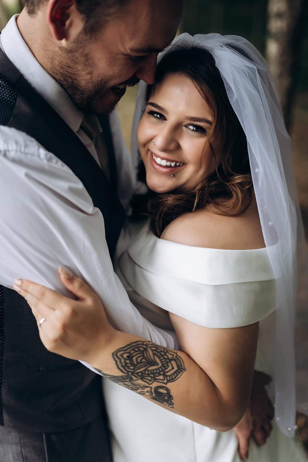 A smiling bride with a tattoo embracing her groom in a tender wedding portrait outdoors.