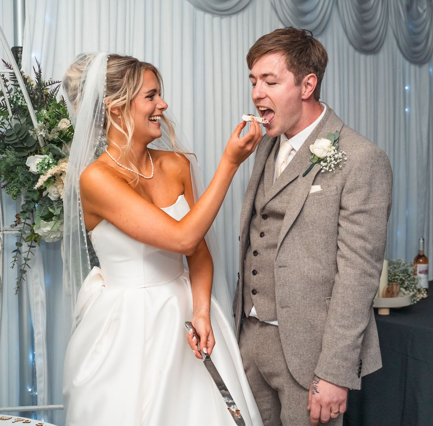 Smiling bride feeding wedding cake to her groom at their elegant white-draped reception venue.