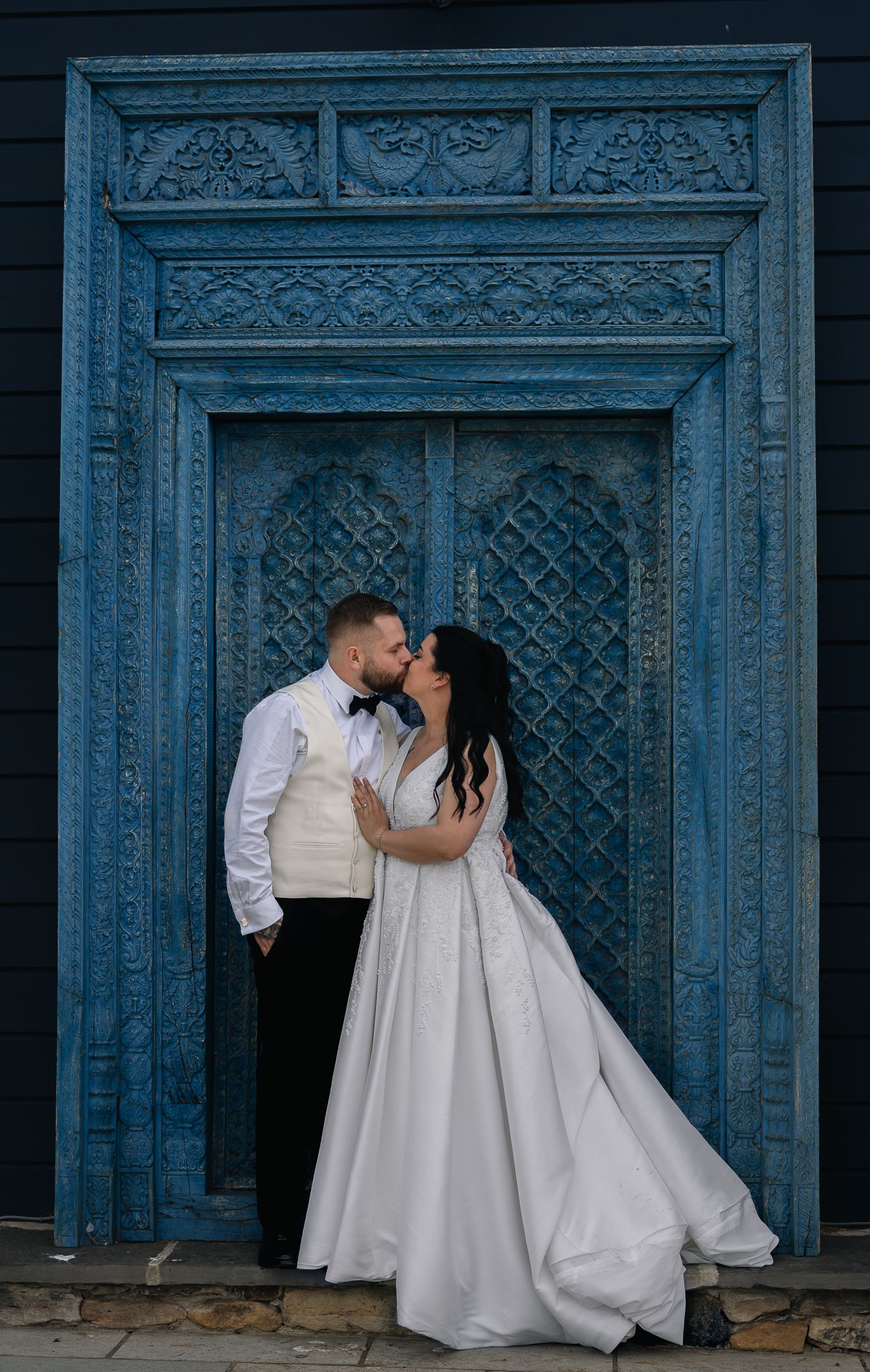 Bride and groom share romantic kiss in front of ornate blue carved wooden doors.