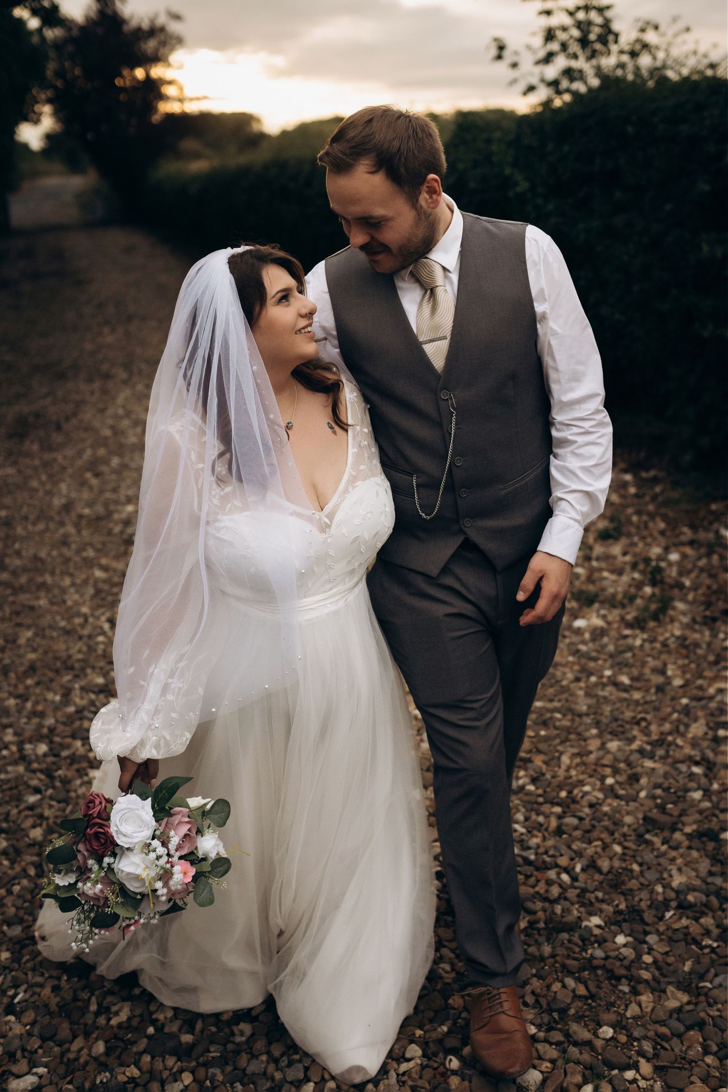A bride and groom walking together at sunset, bride holding a pink and white bouquet in a white gown.