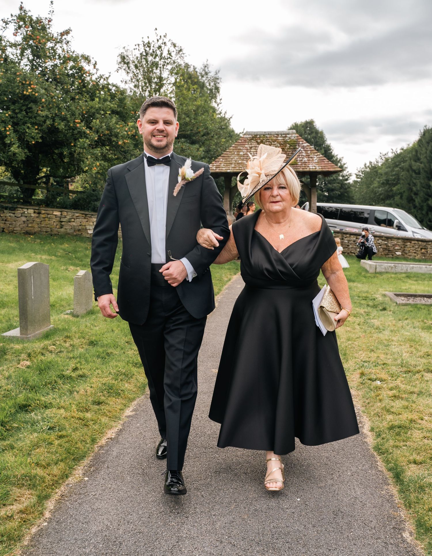 Groom in black tuxedo escorts elegantly dressed woman in black dress at church wedding.