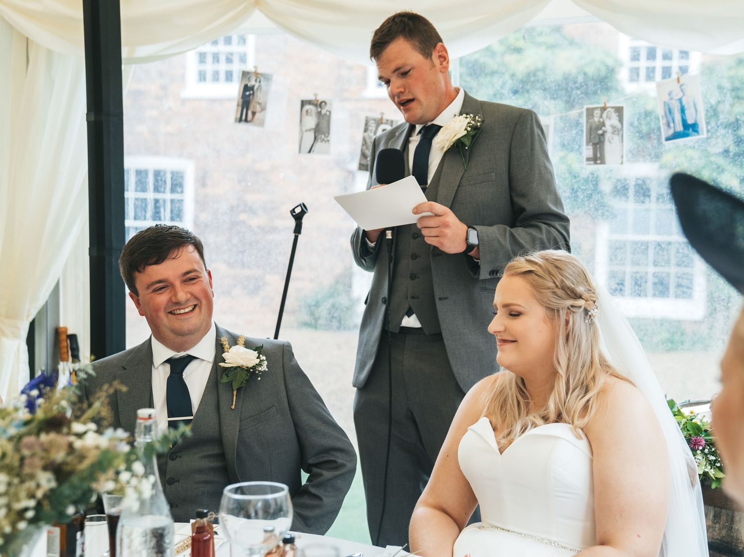 Best man delivering a speech while bride and groom laugh at a wedding reception.