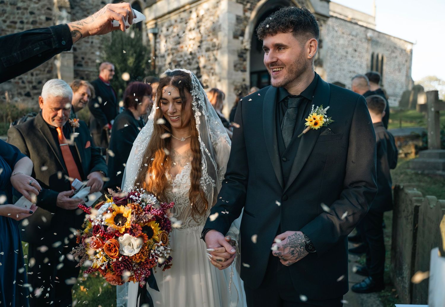 Newlyweds walking through confetti shower outside historic stone church after ceremony.