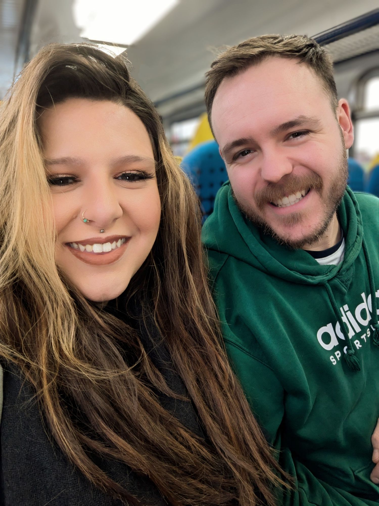 A smiling couple taking a selfie on a train, woman with long blonde hair and man in green Adidas hoodie.