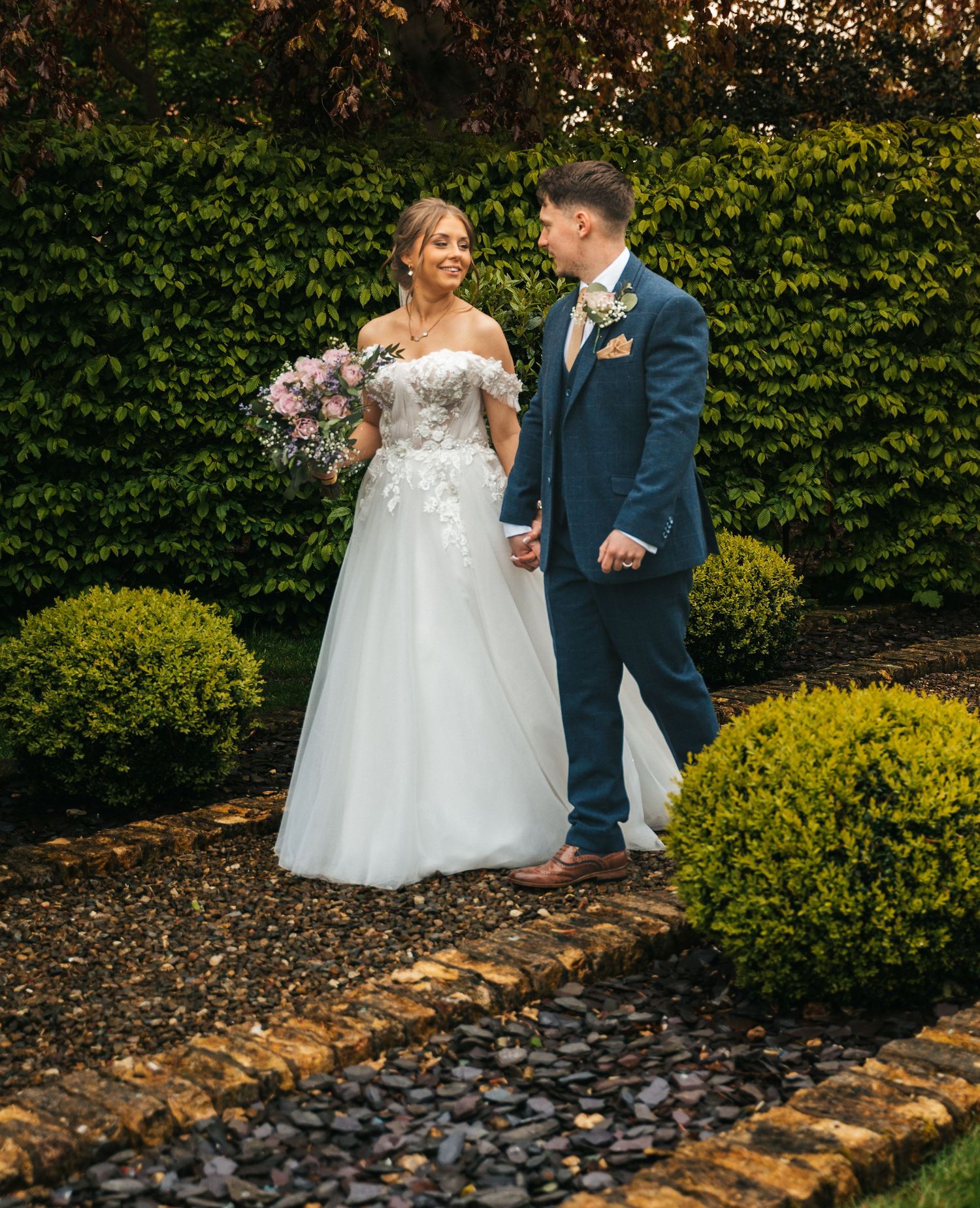 Bride in floral off-shoulder gown and groom in navy suit walking through a lush garden.