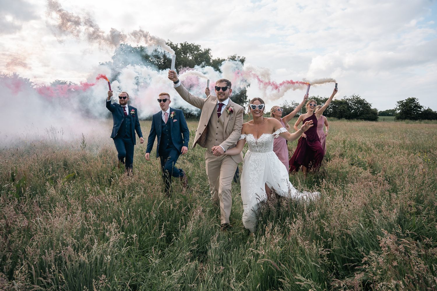 Wedding party runs through field with colorful smoke bombs, bride in white gown, groomsmen in tan suits.