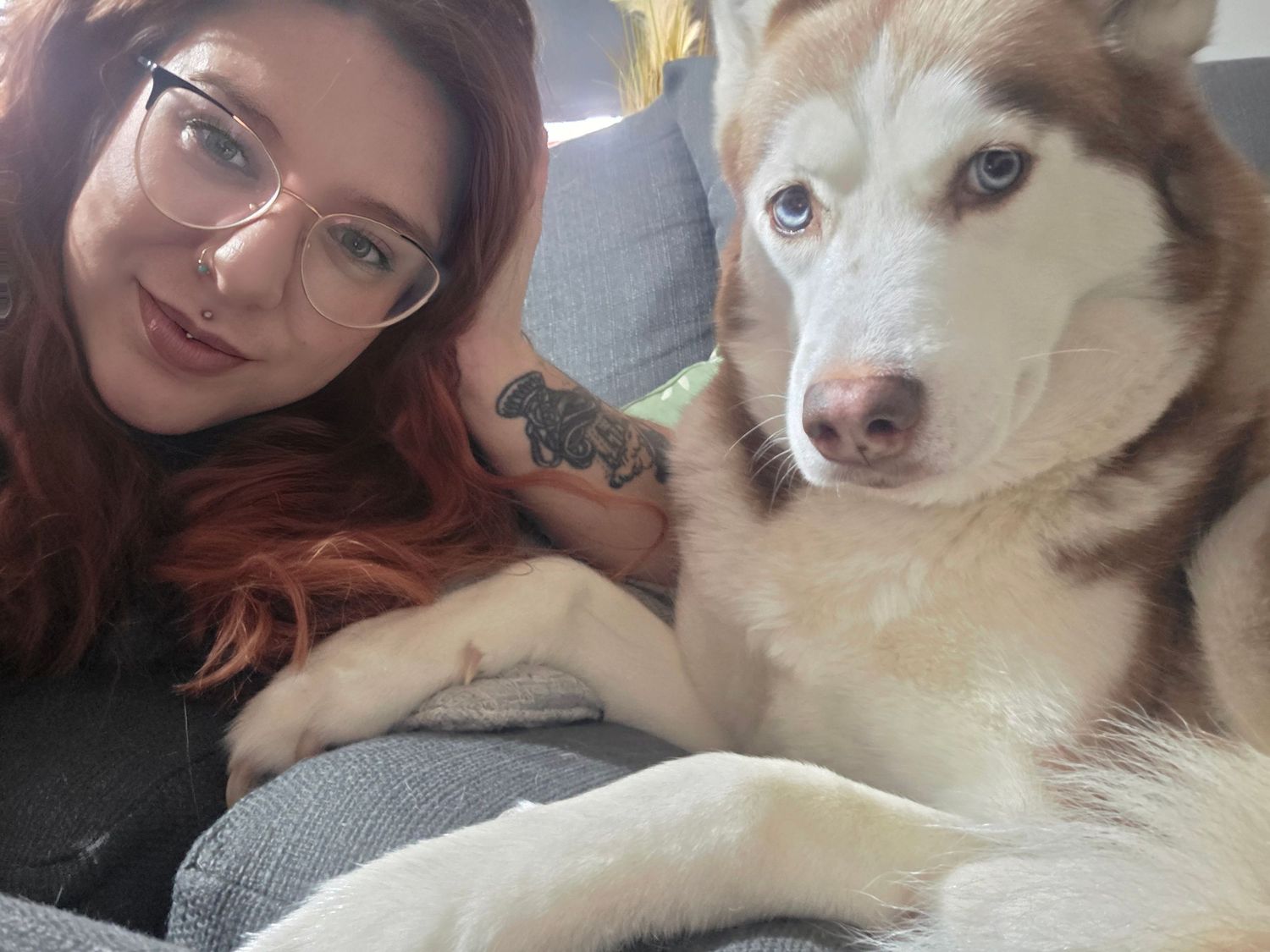 A woman with red hair and glasses posing for a selfie with her Siberian Husky on a couch.
