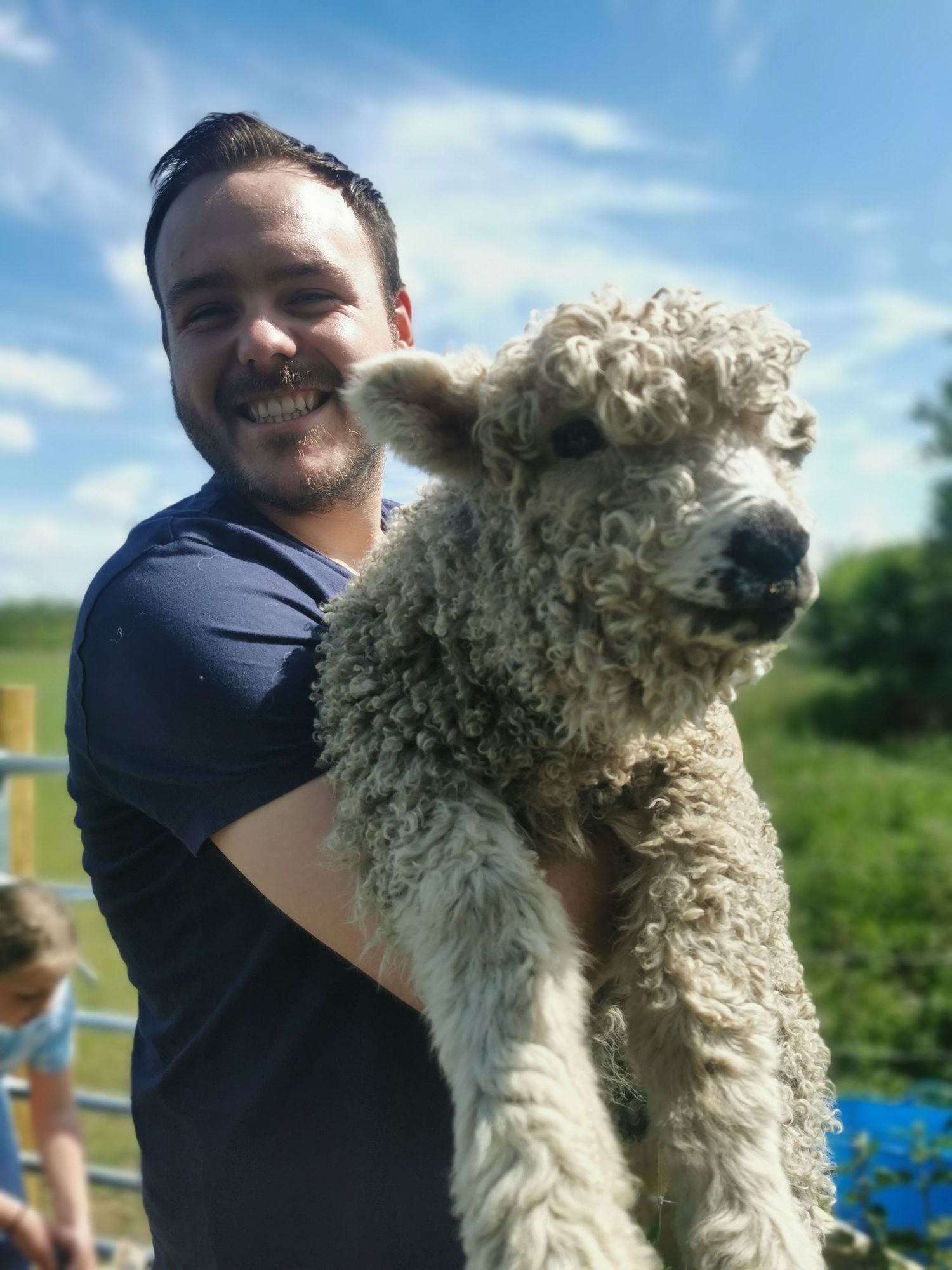 Smiling man in blue t-shirt holding a fluffy white lamb on a sunny farm day.