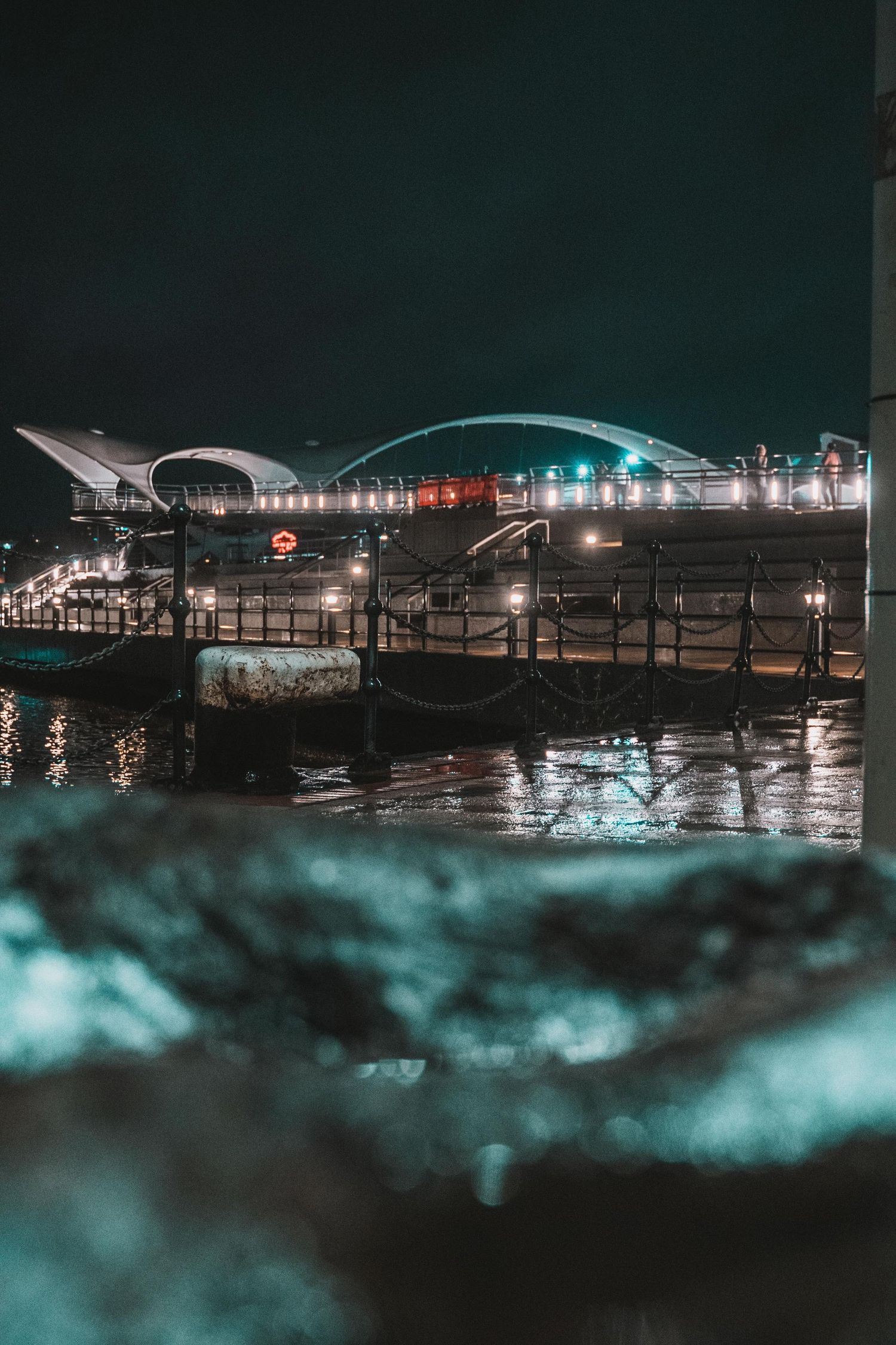 Night cityscape of an illuminated waterfront pier and arched bridge reflecting on calm dark water.