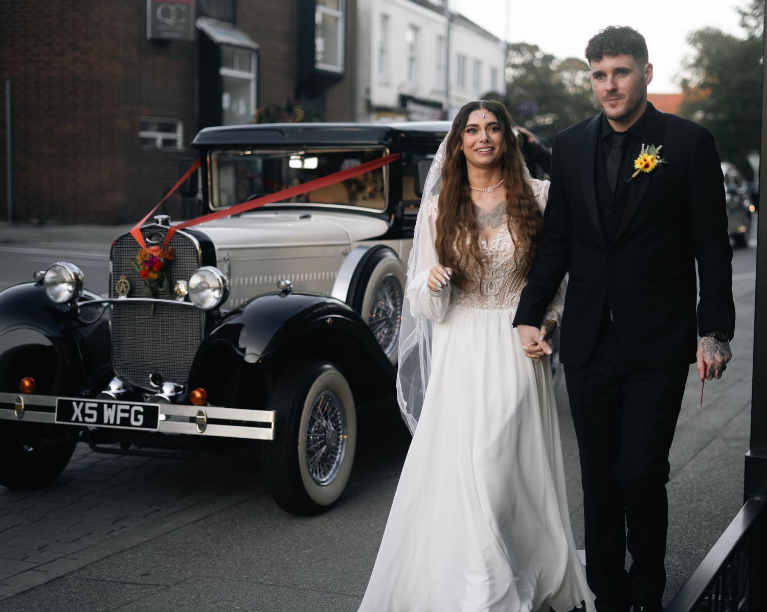 Bride and groom holding hands beside a vintage black wedding car on a street.