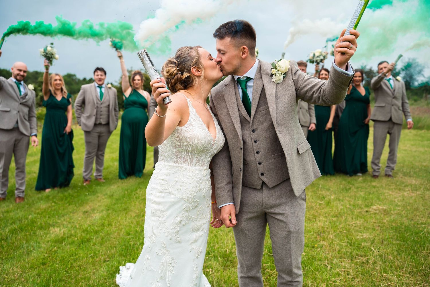 Bride and groom kissing while wedding party celebrates with green smoke bombs outdoors.