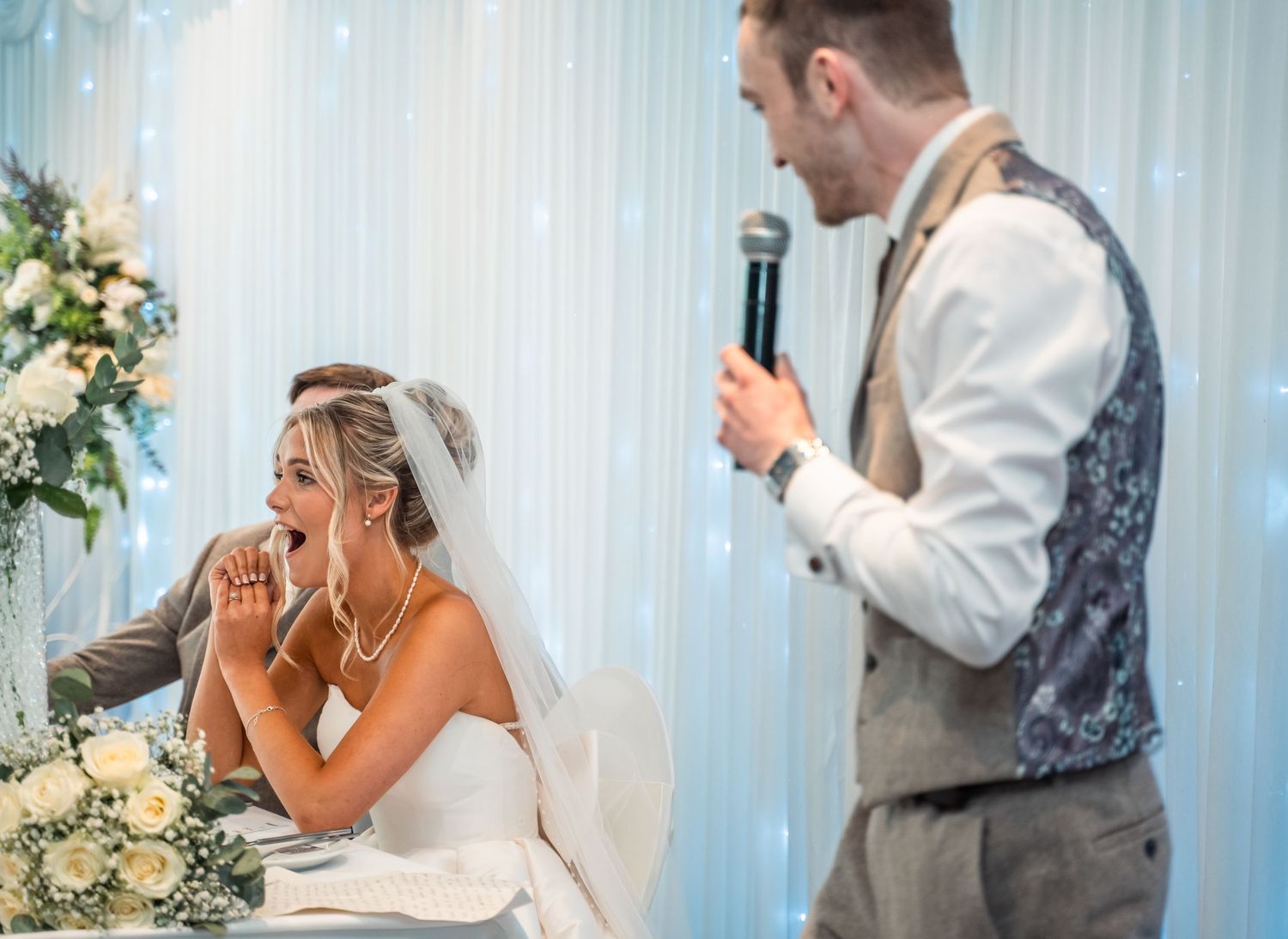 Groom speaks into microphone at wedding reception while bride listens emotionally at table.