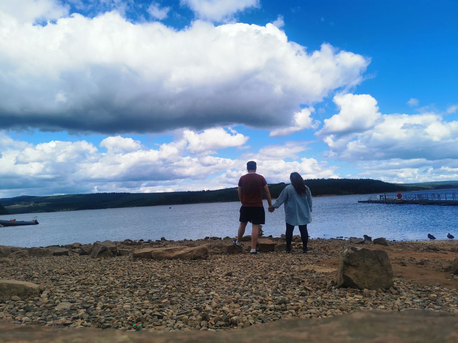 Two people holding hands looking out over a scenic lake under a dramatic cloudy blue sky.