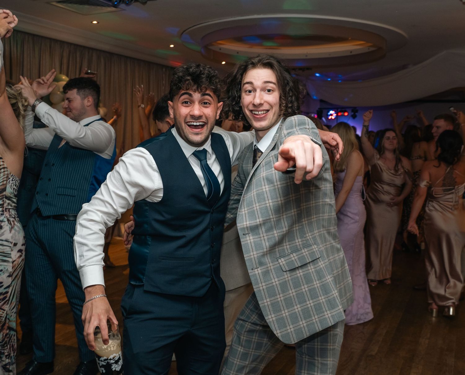 Two cheerful male wedding guests laugh and point at camera on busy dance floor.