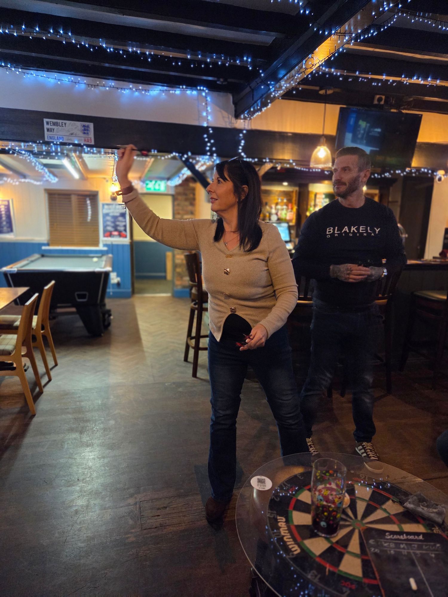 Woman throwing a dart at a dartboard in a pub while a man watches holding a drink.