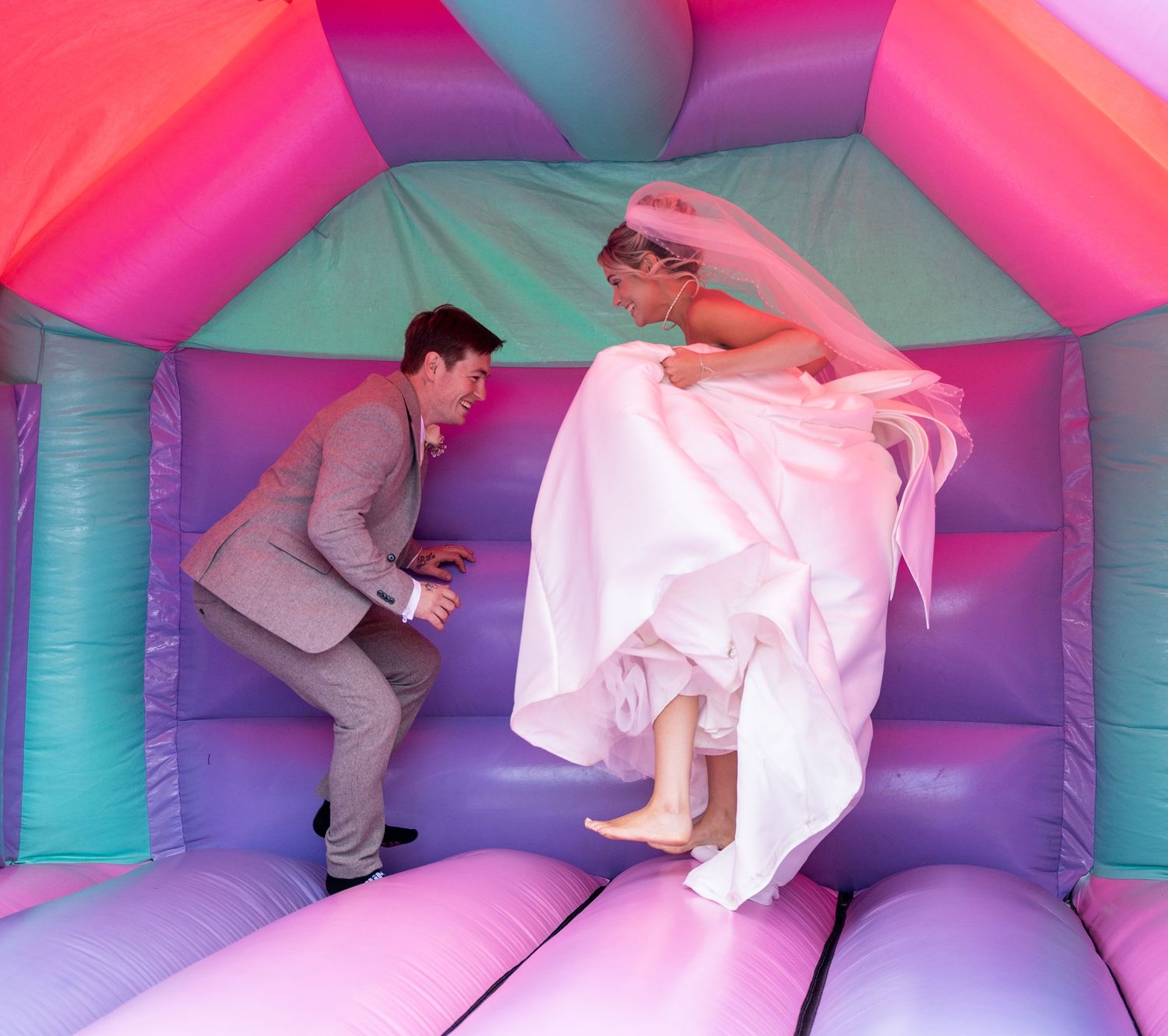 Bride and groom laugh and jump together inside colorful pastel bouncy castle.