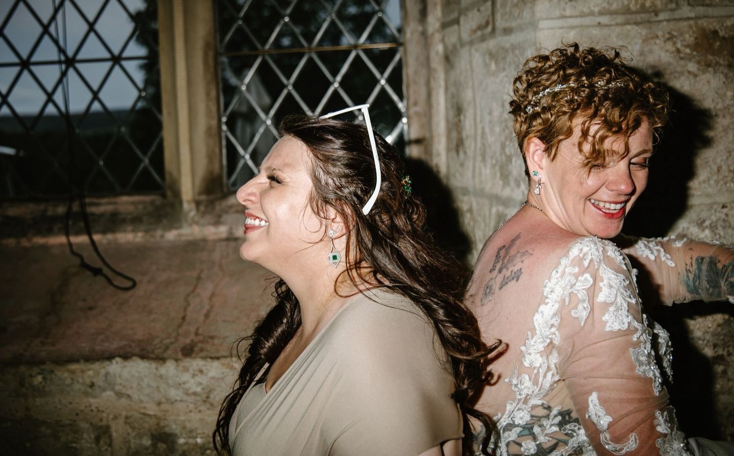 Two women laughing joyfully at a wedding, one wearing a bridal gown with lace sleeves.