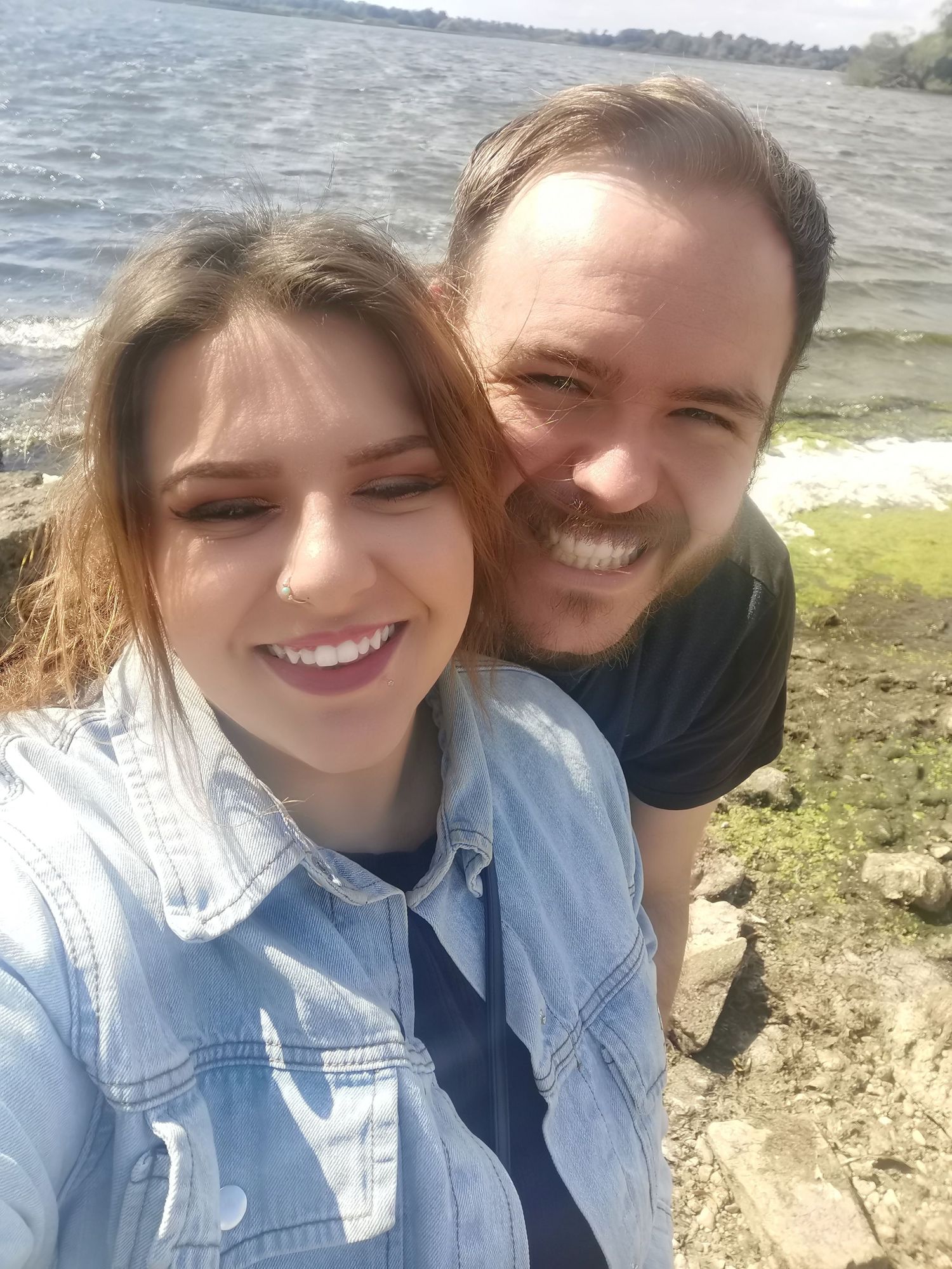 Smiling couple taking a selfie outdoors near a lake on a sunny day.