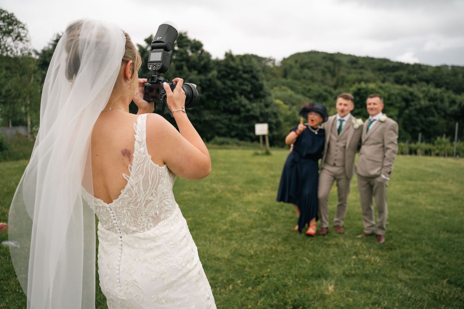 Bride in lace wedding dress photographs guests at outdoor wedding ceremony on green lawn.